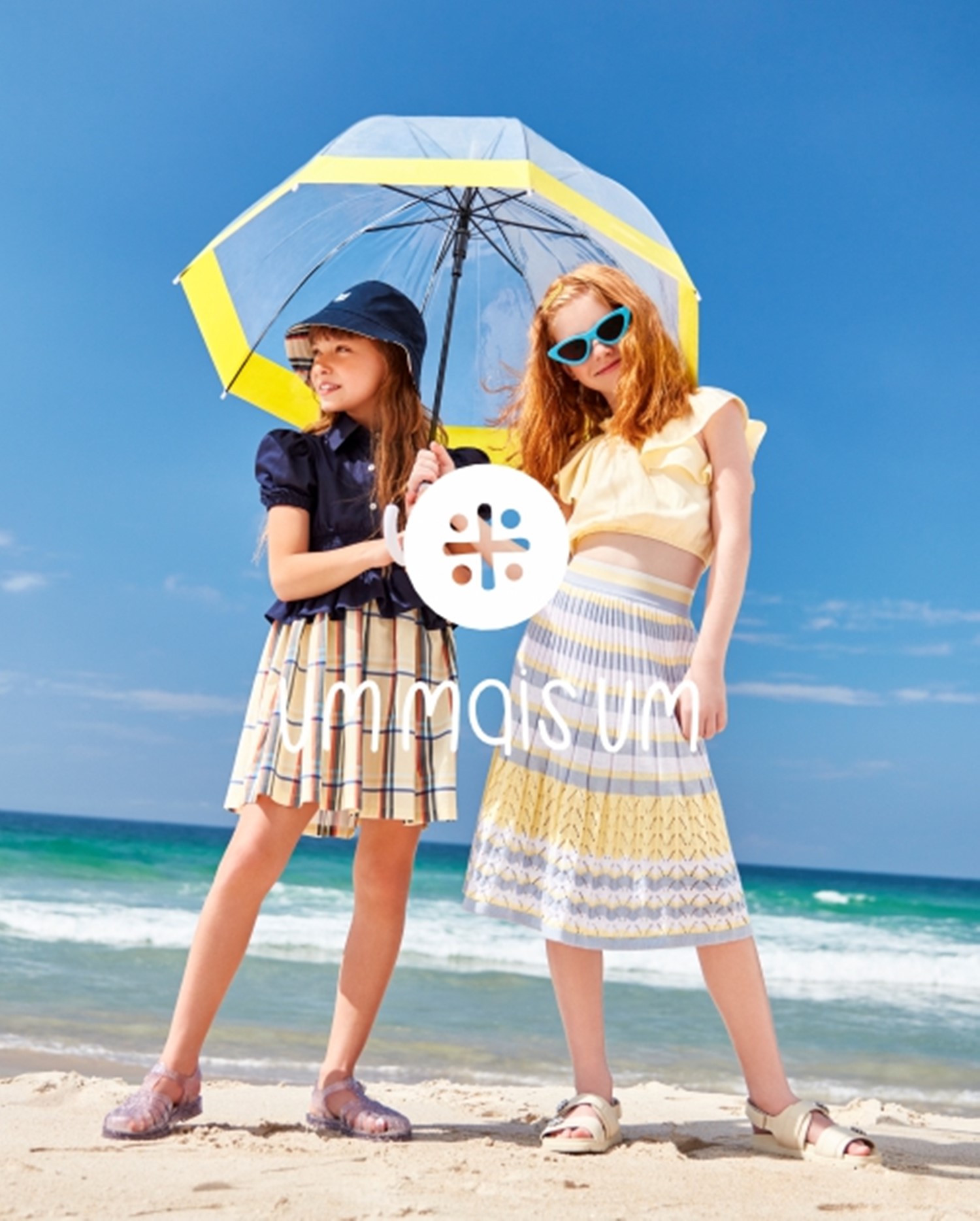 Duas meninas na praia usando looks da Um mais Um e segurando um guarda chuva transparente com borda amarela